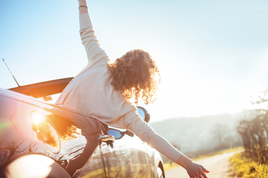 Woman leaning out of a car window with her arm raised, smiling into the sun